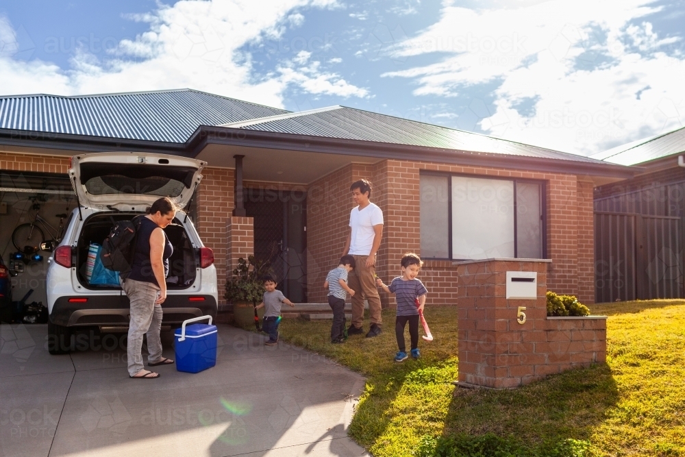 Mum packing family car before sports training with kids playing with hockey sticks on front lawn - Australian Stock Image