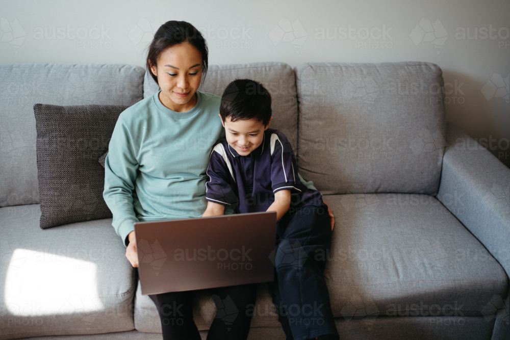 Mum working on her laptop with her son while sitting on the couch - Australian Stock Image