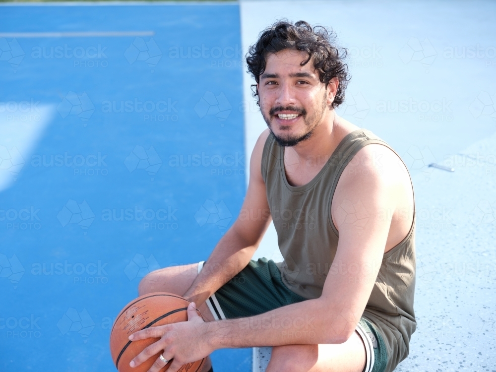 Nepalese man sitting on a basketball court smiling and holding a basketball - Australian Stock Image