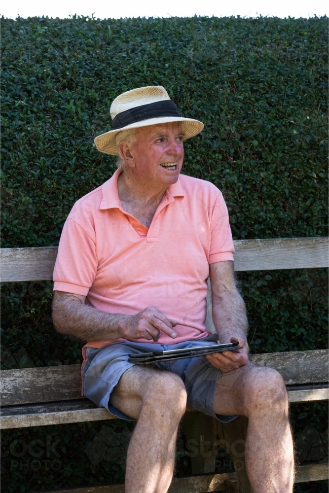 Old man sitting on a bench outside with his tablet - Australian Stock Image