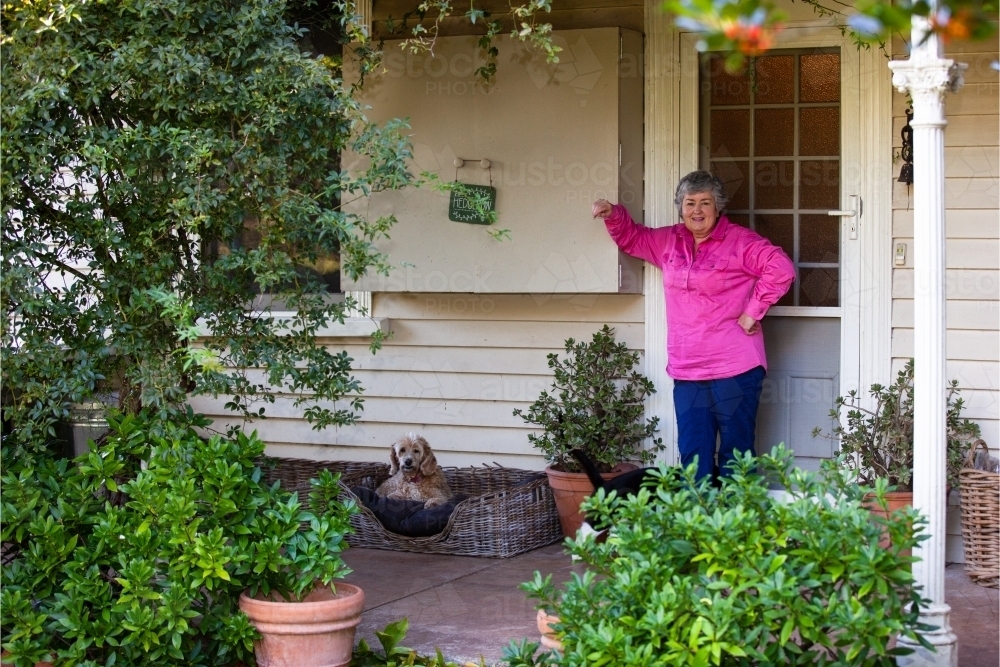 Old woman in pink coat standing outside the door with her pet dog - Australian Stock Image