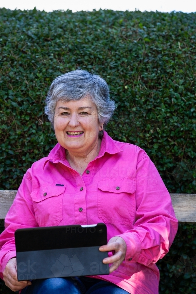 Old woman sitting on the bench smiling while using her tablet - Australian Stock Image