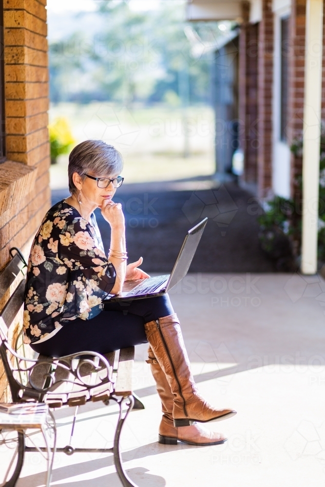 Older woman with laptop on sunlit verandah of home working through covid-19 - Australian Stock Image