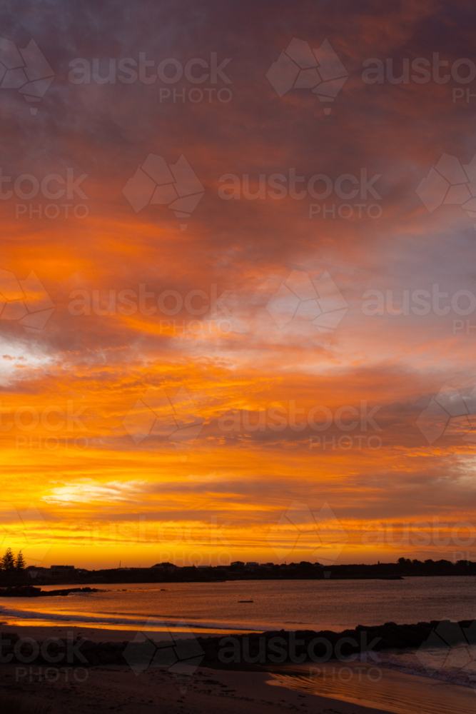 Orange, dramatic sunset over beach and coastal town - Australian Stock Image
