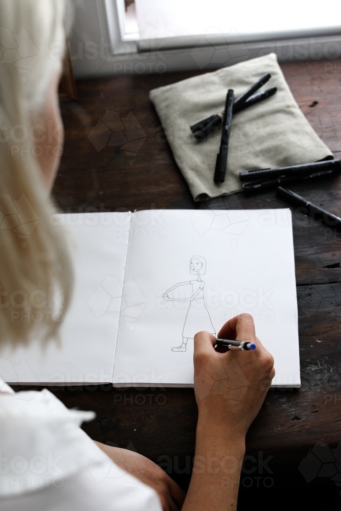 Over the shoulder perspective of a young blonde woman drawing in a sketchbook - Australian Stock Image