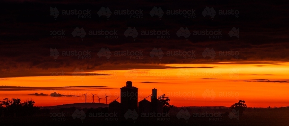 Panoramic view of a red sky during dawn with the silhouettes of silos and wind turbines - Australian Stock Image