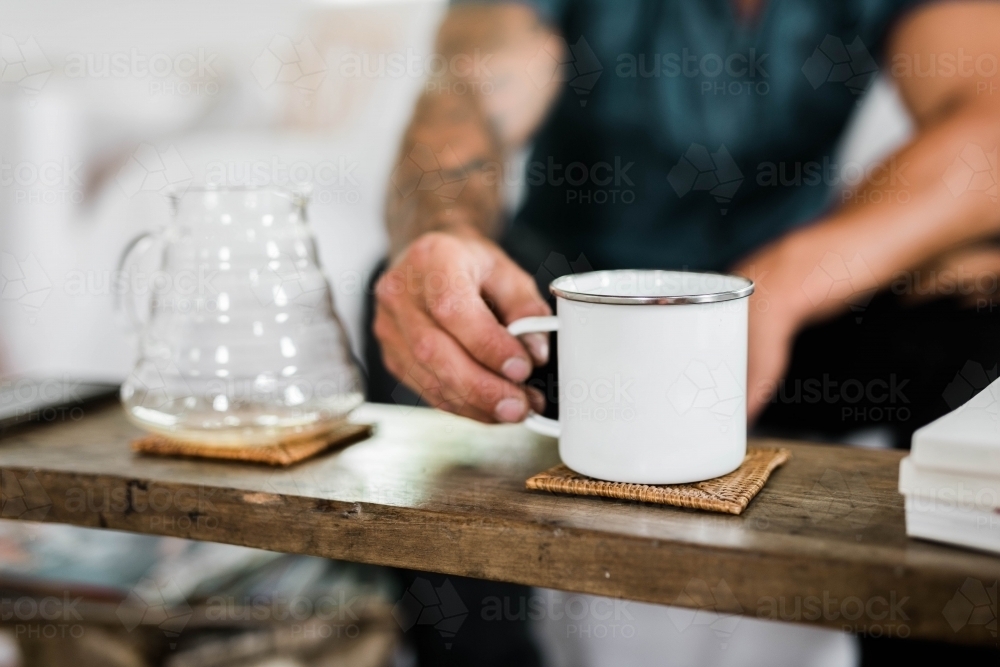 Person picking up a cup of coffee next to a pour over vessel - Australian Stock Image