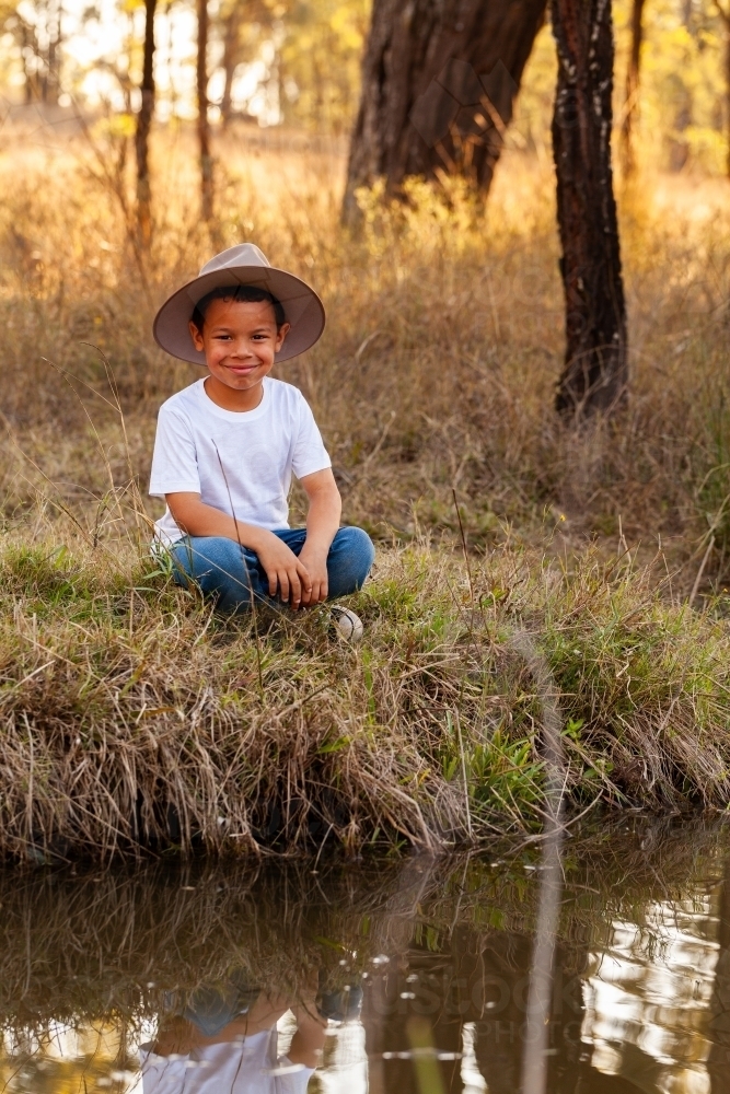 Portrait of a first nations Australian boy sitting by creek in paddock - Australian Stock Image