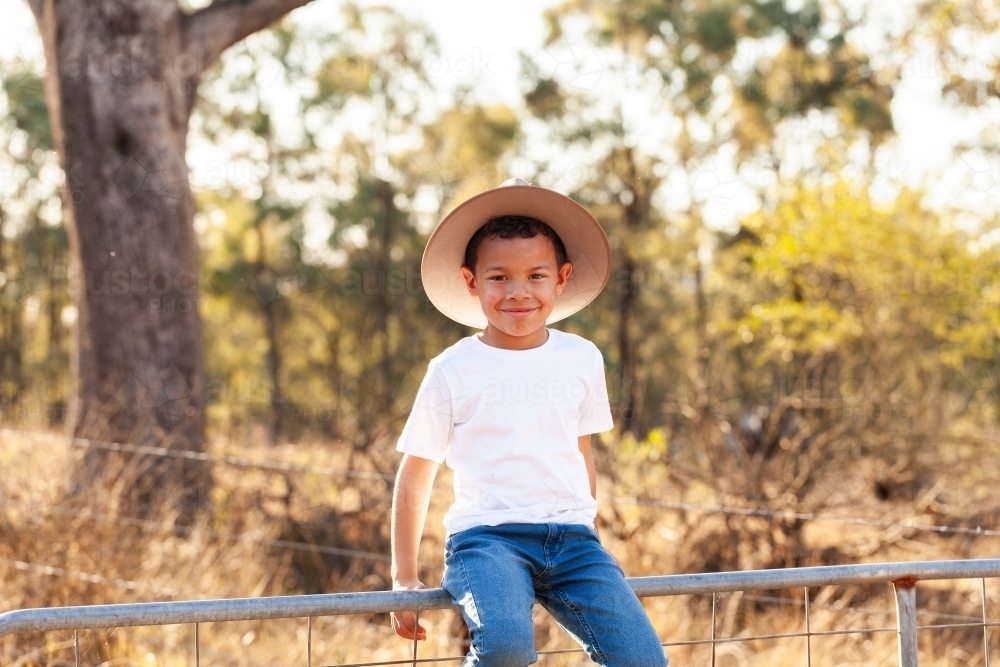 Portrait of a first nations Australian boy smiling and sitting on gate in a paddock - Australian Stock Image