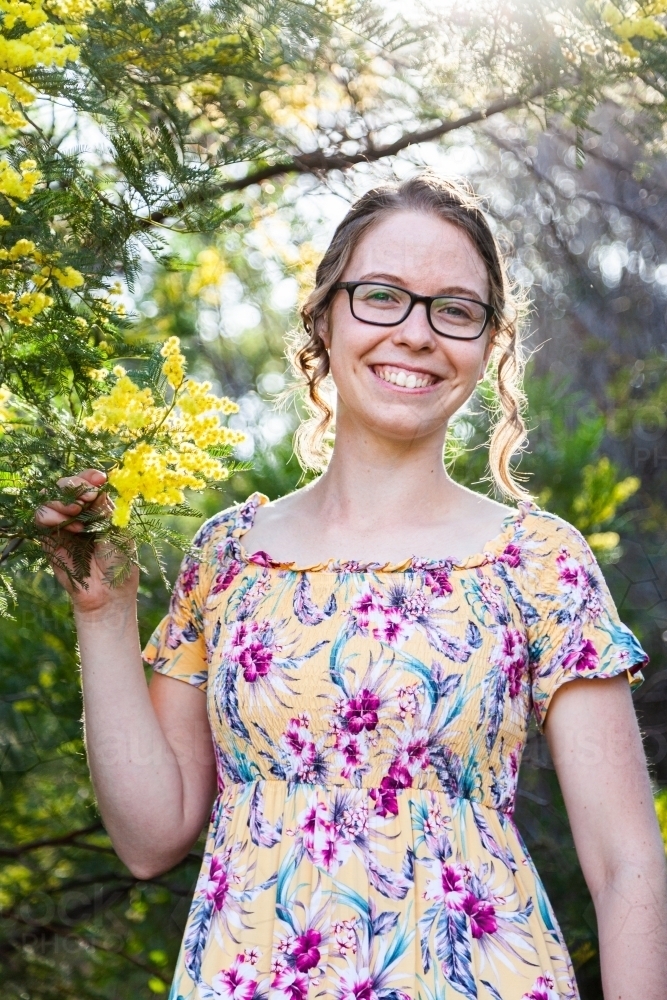 Portrait of a happy smiling young woman with glasses standing among leaves and blossoms of wattle - Australian Stock Image