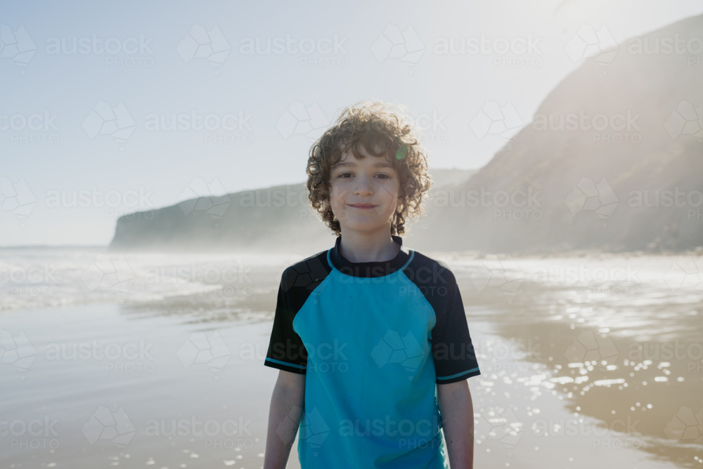 Portrait of a happy young boy with curly hair at the beach in the afternoon looking at the camera - Australian Stock Image