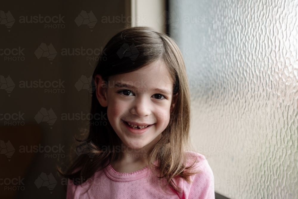 Portrait of a happy young girl smiling with a missing front tooth sitting by a window - Australian Stock Image