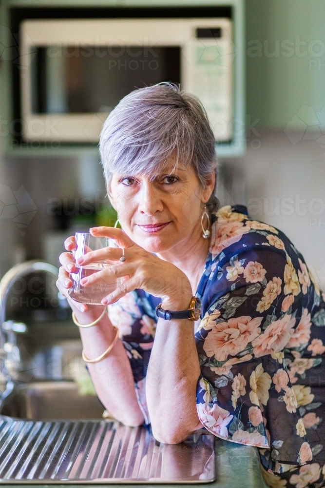 Portrait of a healthy senior woman leaning on the kitchen bench drinking a glass of water - Australian Stock Image