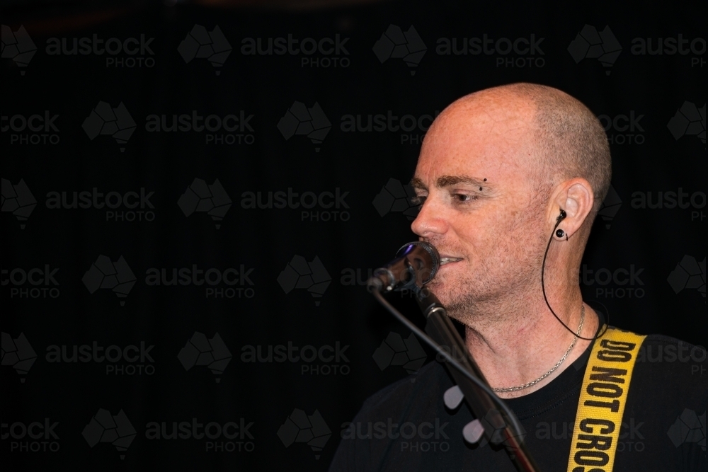 Portrait of a male entertainer singing at microphone with shaved head and black background - Australian Stock Image
