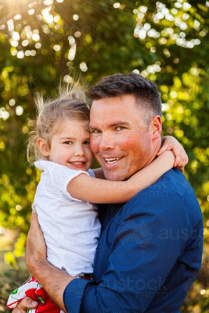Portrait of a smiling father hugging his happy daughter in golden sunlight - Australian Stock Image