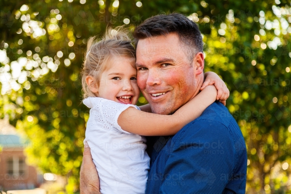 Portrait of a smiling father hugging his happy daughter in golden sunlight - Australian Stock Image