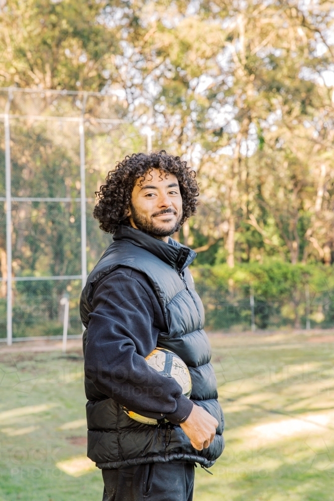 Portrait of a smiling man on his side with a soccer ball on his hand, standing on the field - Australian Stock Image