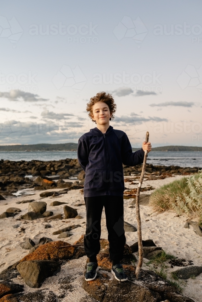 Portrait of a young boy posing with a walking stick on a beach at sunset - Australian Stock Image