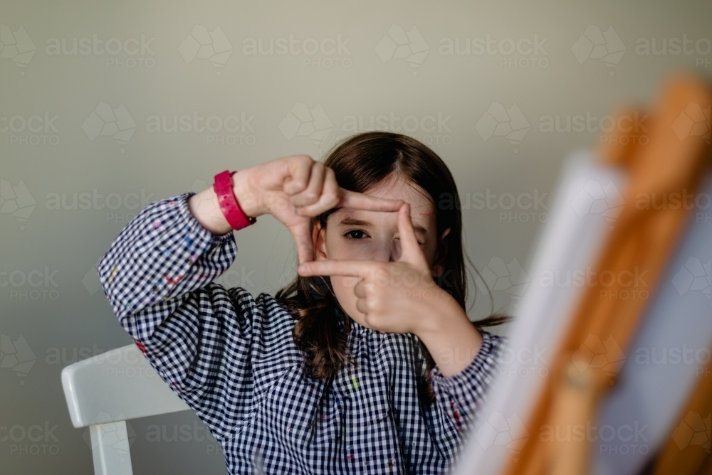 Portrait of a young girl at a painting easel, looking through hands forming a finger frame - Australian Stock Image