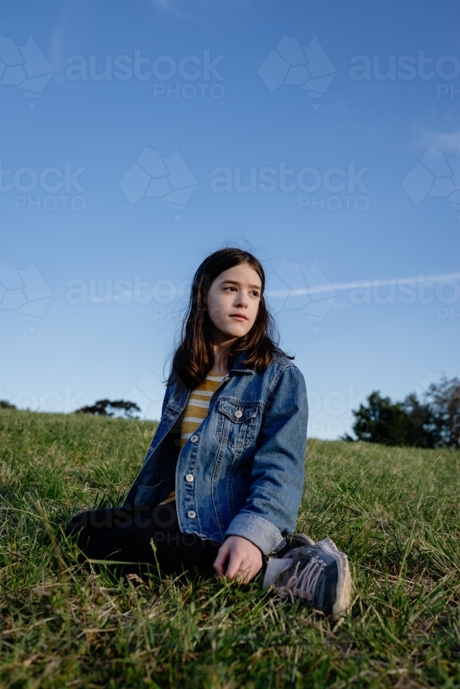 Portrait of a young girl wearing a denim jacket sitting on a grassy hill against a blue sky - Australian Stock Image