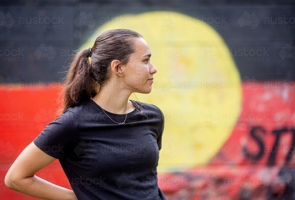Portrait of an Aboriginal woman standing with a red, yellow and black flag mural in the background - Australian Stock Image