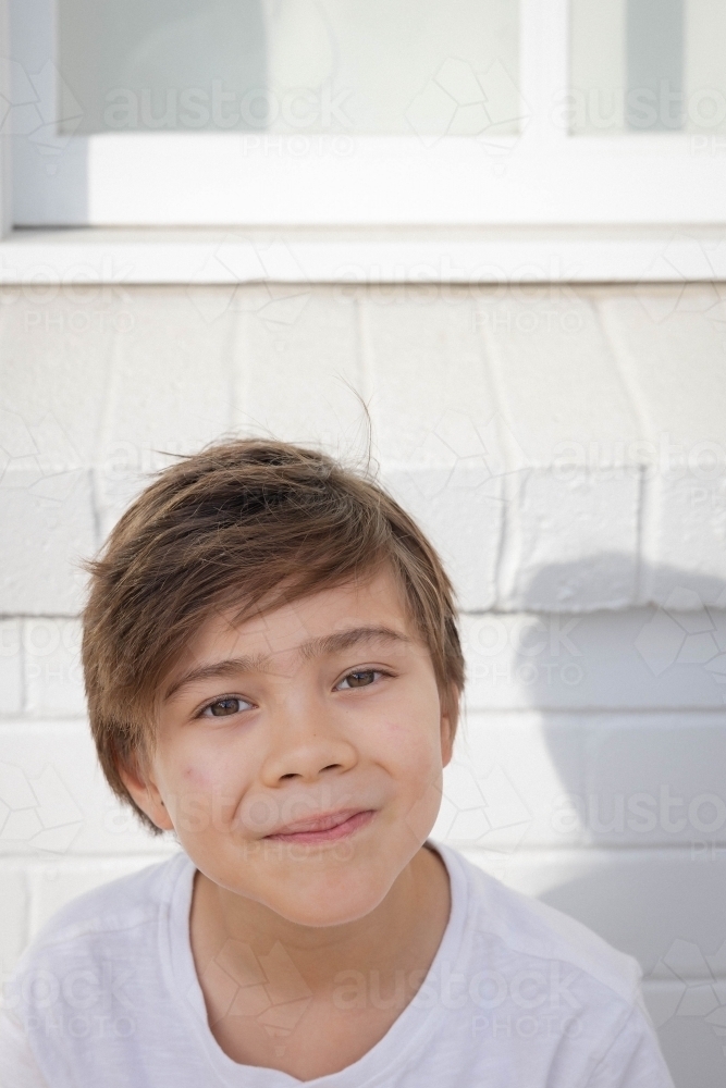 Portrait of boy sitting against white brick wall - Australian Stock Image