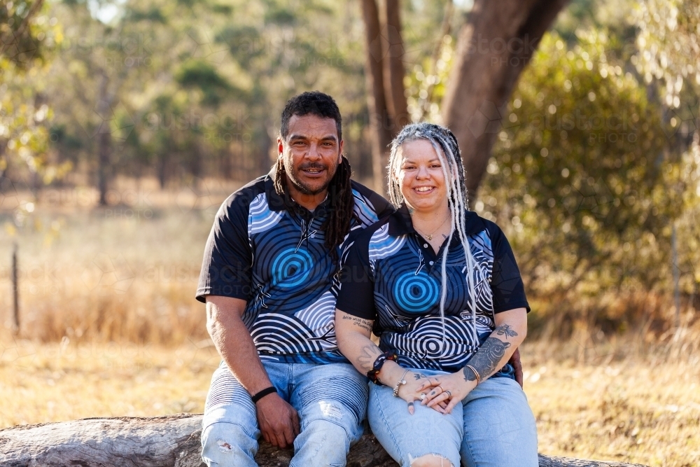 Portrait of happy aboriginal couple sitting together on log - Australian Stock Image