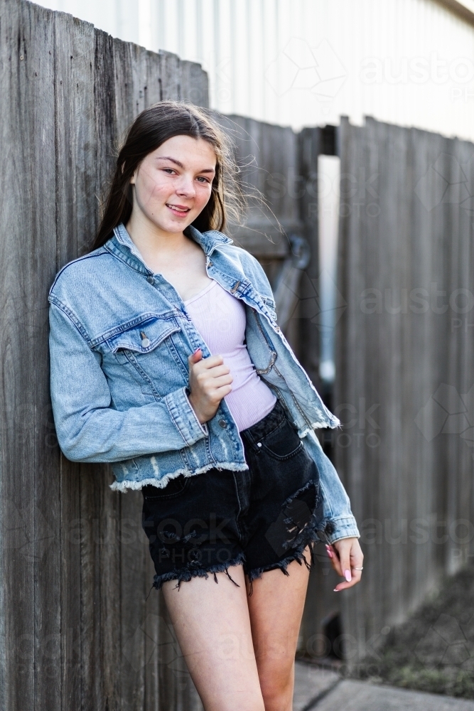 Portrait of happy teenage girl leaning on wooden backyard fence after school - Australian Stock Image
