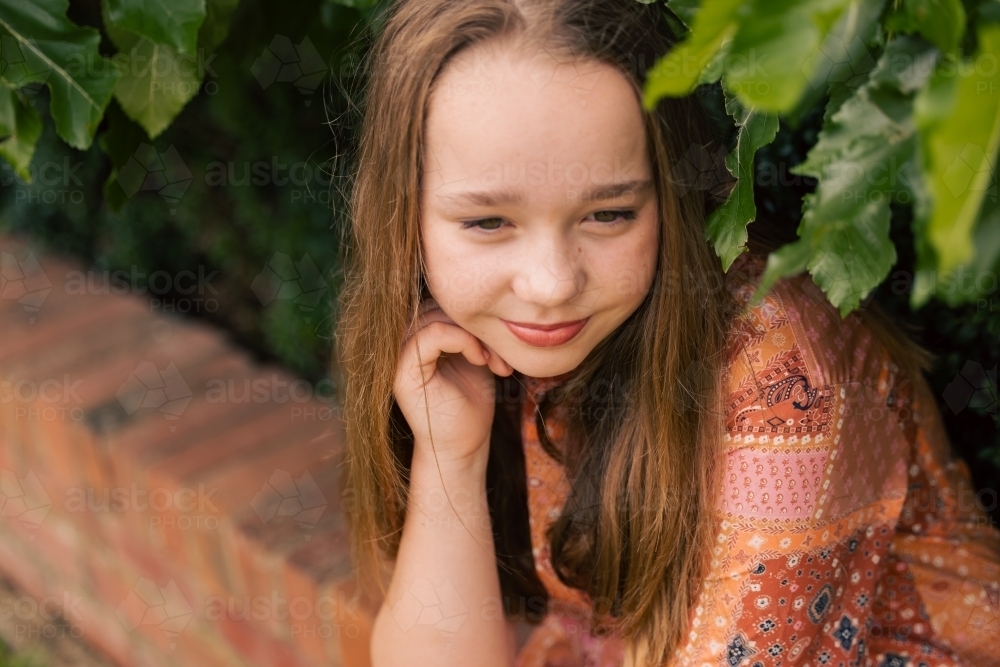 Portrait of pre-teen girl sitting on brick wall with natural green leafy background - Australian Stock Image