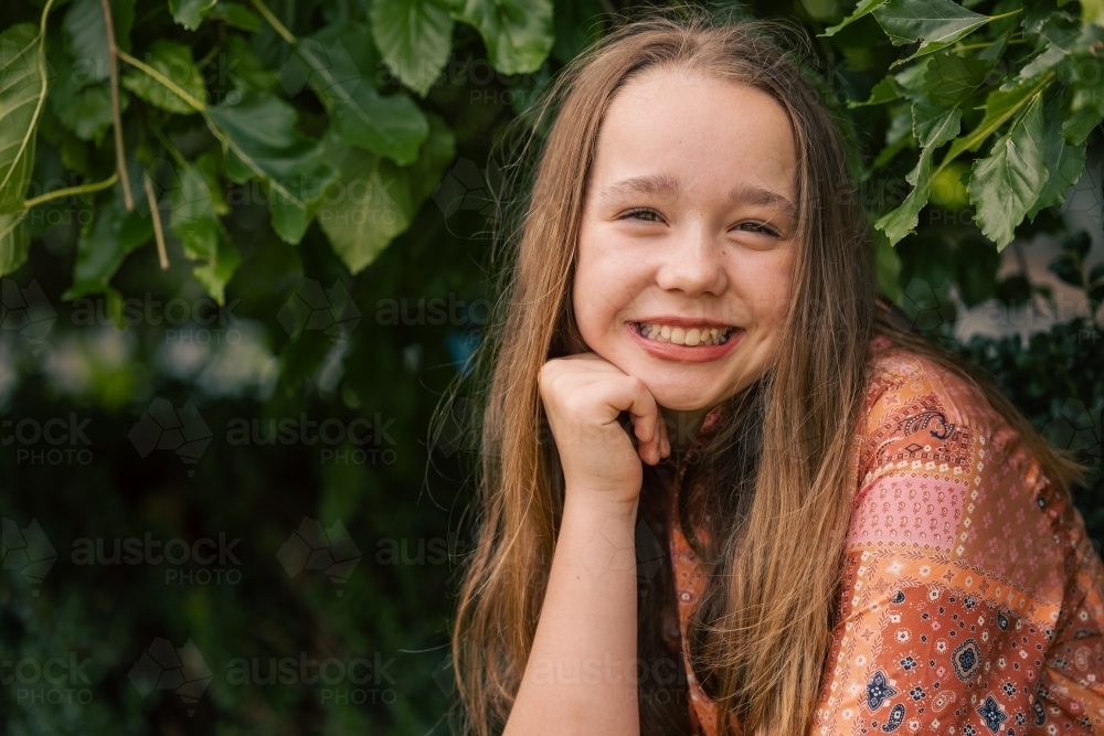 Portrait of pre-teen girl sitting on brick wall with natural green leafy background - Australian Stock Image