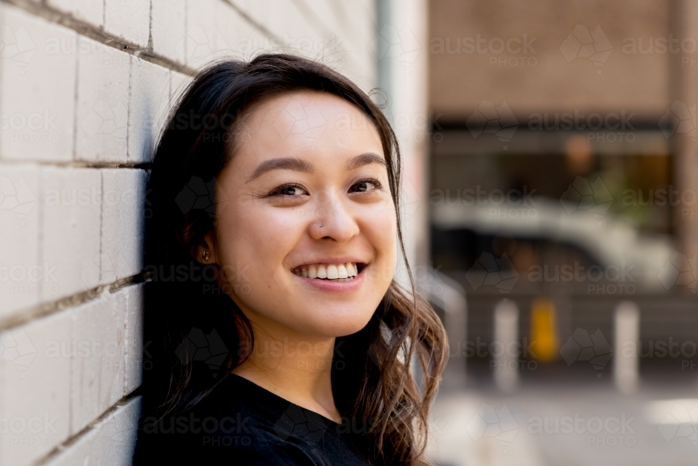 portrait of smiling Asian woman, casual portrait leaning against a brick wall - Australian Stock Image