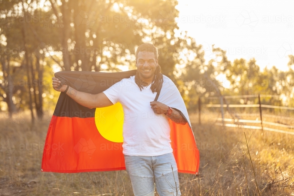 Portrait of smiling first nation australian man holding aboriginal flag in paddock - Australian Stock Image