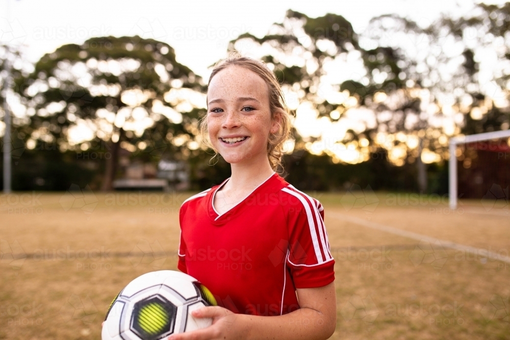 Portrait of tween girl in a red uniform holding a football and smiling - Australian Stock Image