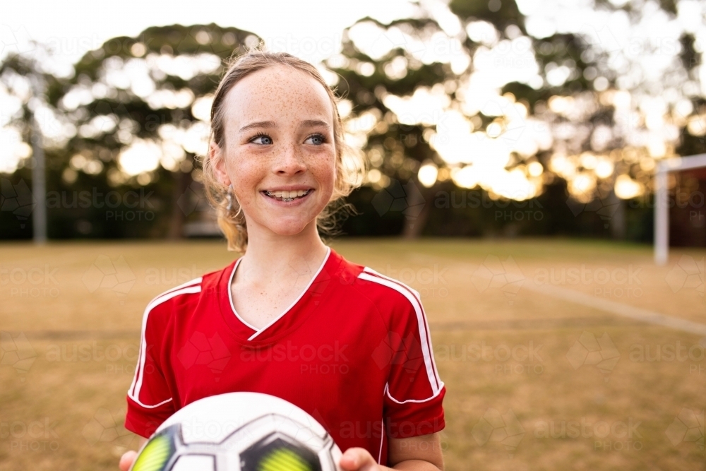 Portrait of tween girl in a red uniform holding a football and smiling - Australian Stock Image