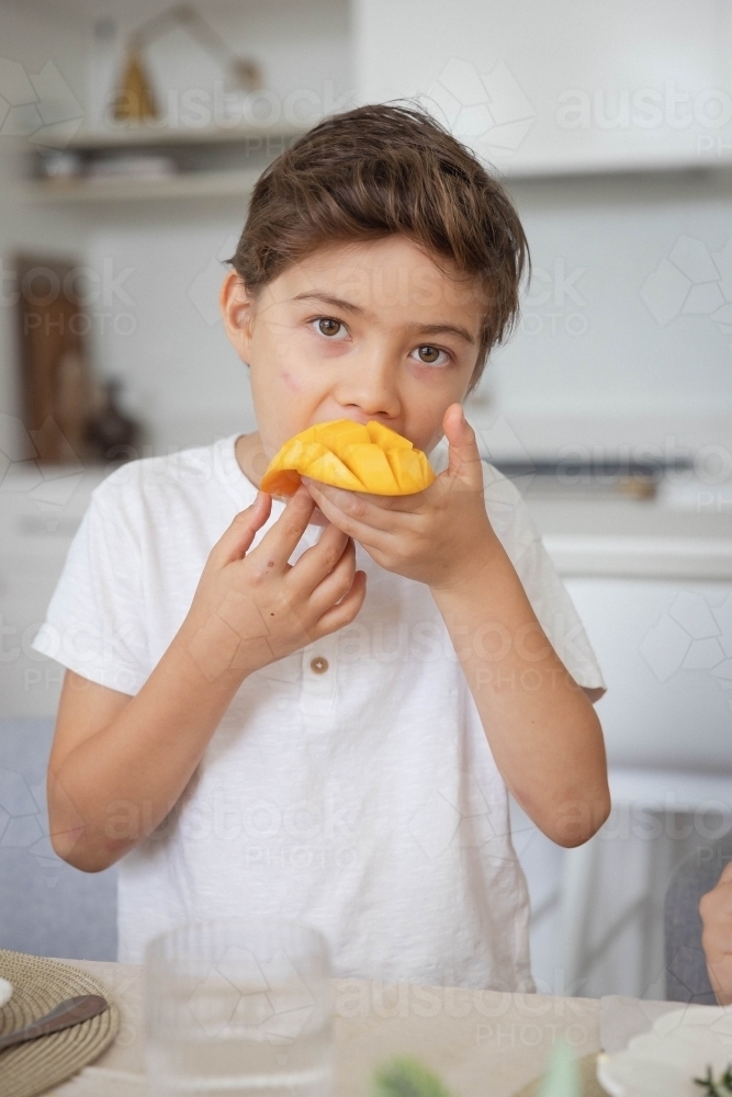 Portrait of young boy eating slice of mango in kitchen - Australian Stock Image