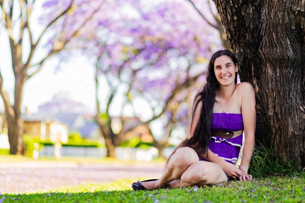 Portrait of young woman in her late twenties relaxing, sitting beside a tree in a park - Australian Stock Image
