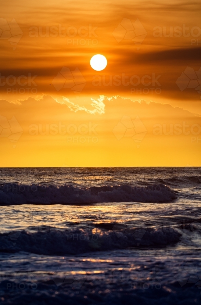 Portrait view of the orange sun rises on the ocean while the waves roll in. - Australian Stock Image