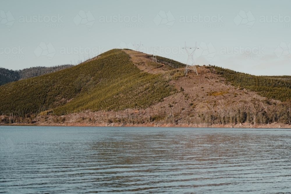 Powerlines on a mountain through a pine forest clearing with a lake in the foreground - Australian Stock Image