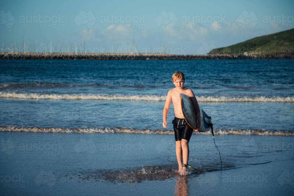 Pre-teen boy carrying surf board out of the water on the beach at Coffs Harbour - Australian Stock Image