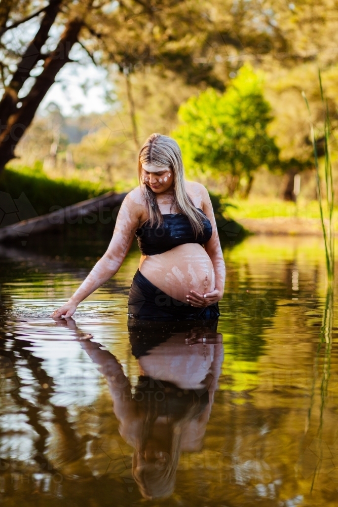 Pregnant Aboriginal Australian woman in traditional ochre body paint standing in creek - Australian Stock Image