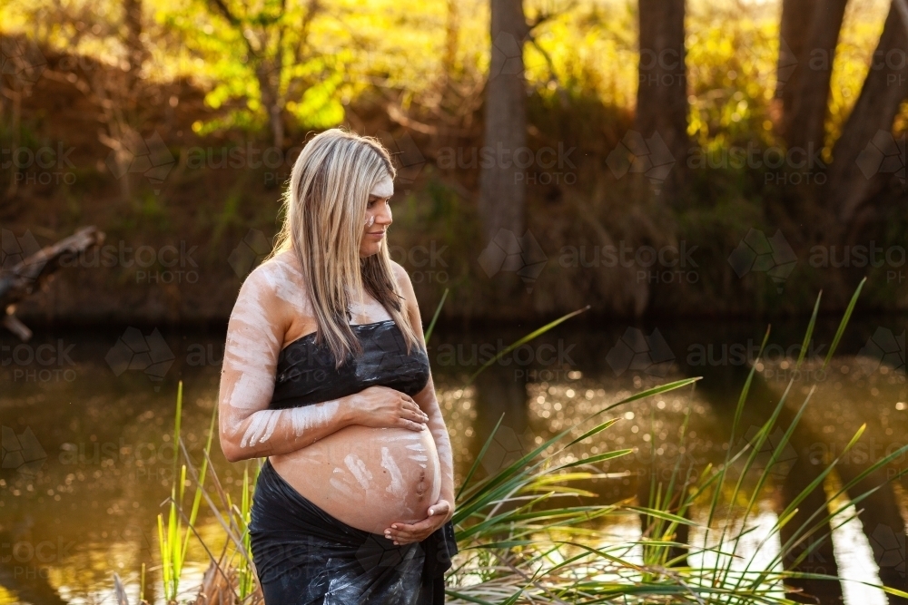 Pregnant Aboriginal Australian woman standing by creek on summer afternoon - Australian Stock Image