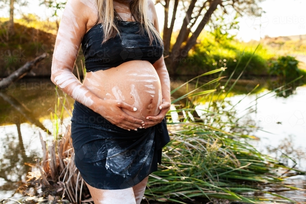 Pregnant Aboriginal Australian woman standing by creek on summer afternoon - Australian Stock Image