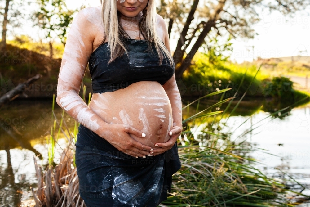 Pregnant Aboriginal Australian woman standing by creek on summer afternoon - Australian Stock Image