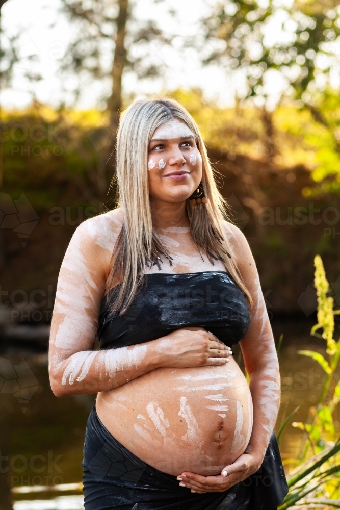 Pregnant Aboriginal Australian woman standing by creek on summer afternoon - Australian Stock Image