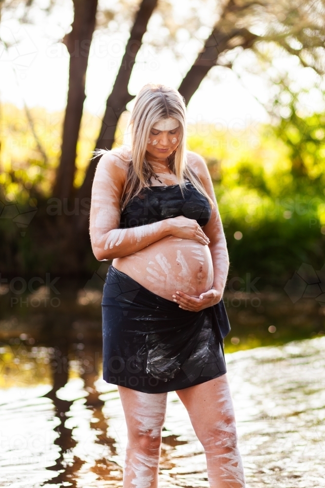 Pregnant Aboriginal lady in ochre body paint standing by water in bushland - Australian Stock Image