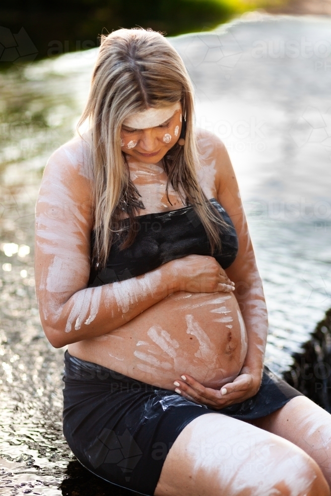 Pregnant Aboriginal lady in traditional ochre body paint sitting in creek water in bushland - Australian Stock Image