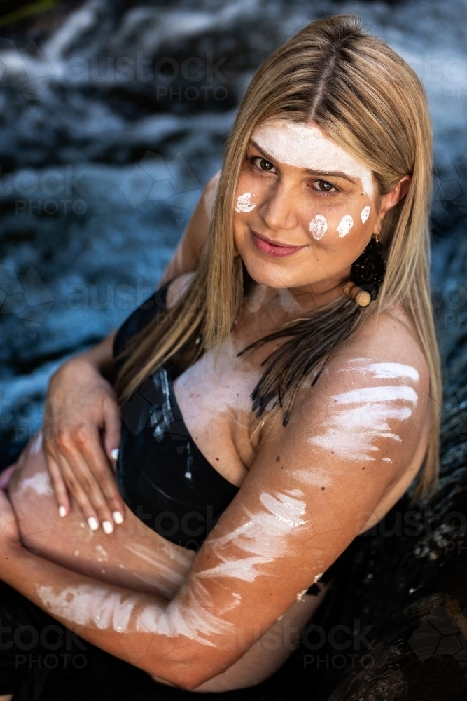 Pregnant Aboriginal lady in traditional ochre body paint sitting in waterfall in bushland - Australian Stock Image