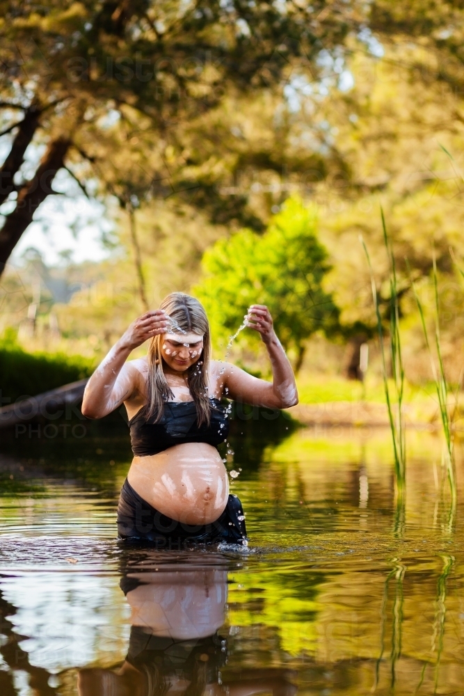 Pregnant Aboriginal woman in traditional body paint splashing streams of water droplets in creek - Australian Stock Image