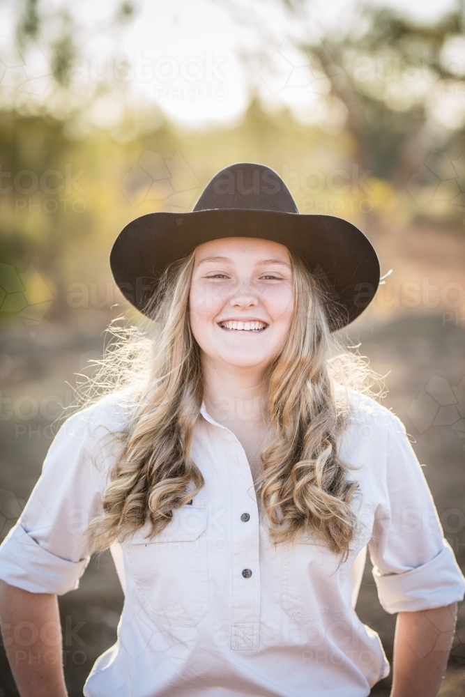 Pretty teenage girl standing on farm in drought smiling wearing akubra hat - Australian Stock Image