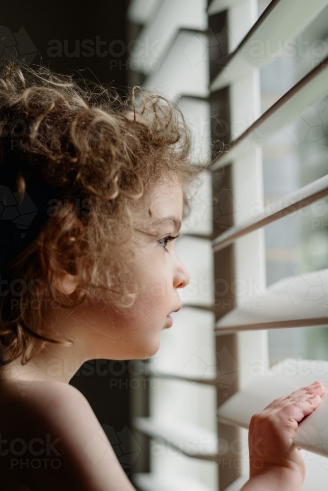 Profile of a young toddler boy with curly hair looking out louvre window - Australian Stock Image